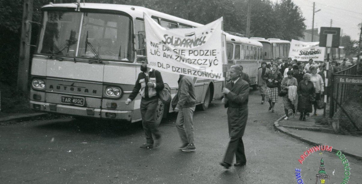 Archwialne zdjęcie ze stanu wojennego, ludzie idący w kolumnie, niosący napisy Solidarność, w tle kolumna autobusów