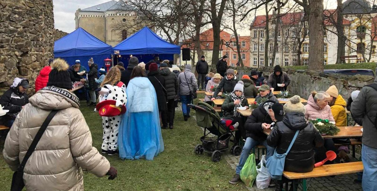 Ludzie siedzący przy stołach, po bokach kamienne, zabytkowe mury obronne