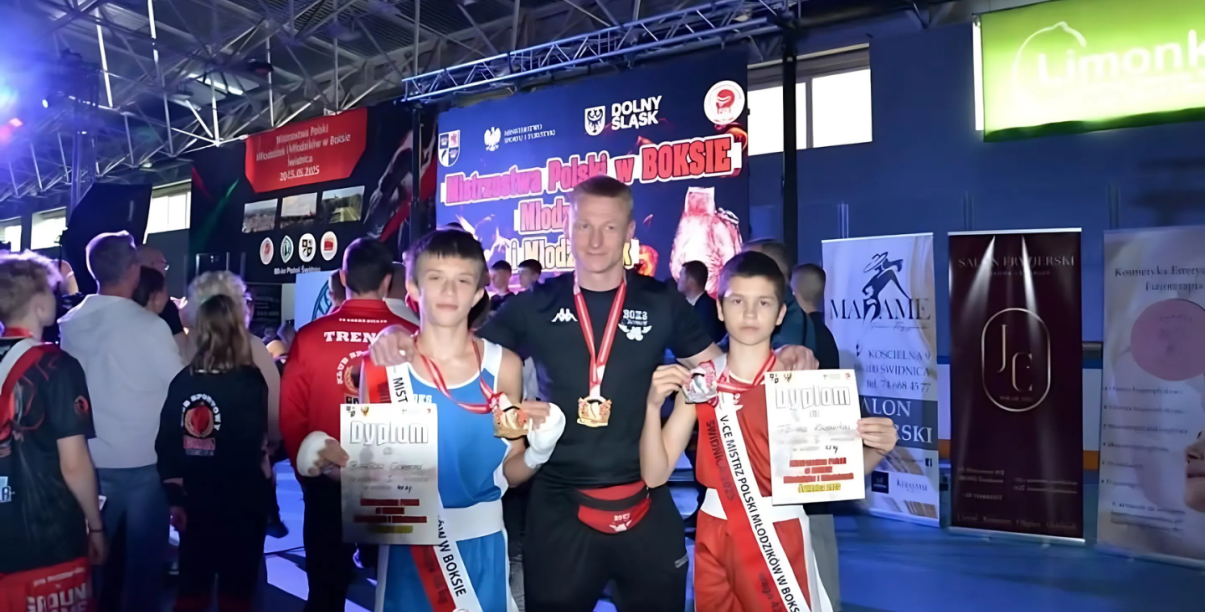 Dwóch młodych zawodników w sportowych strojach, z dyplomami i medalami, pomiędzy nimi trener
