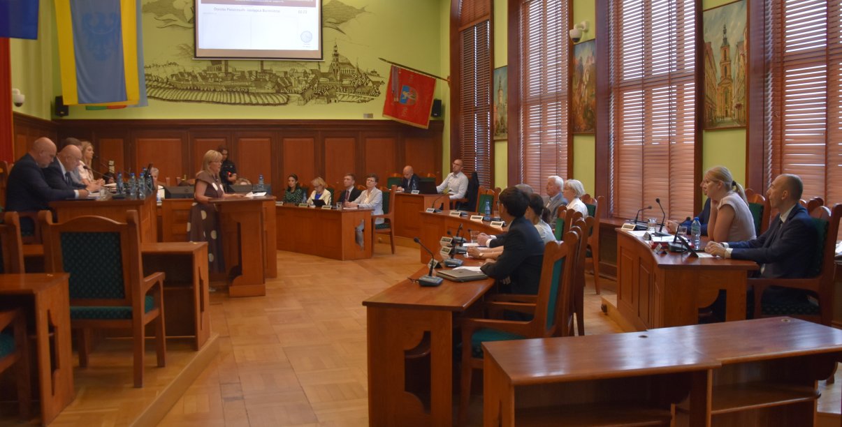 Duża sala z parkietem i drewnianymi stołami, przy stołąch siedzą radni