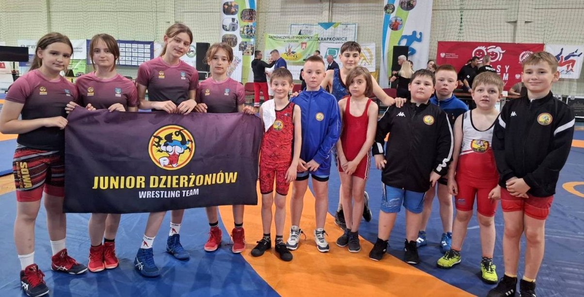 Grupowe zdjęćie kilkunastu młodych zapaśniczek i zapaśników w hali sportowej