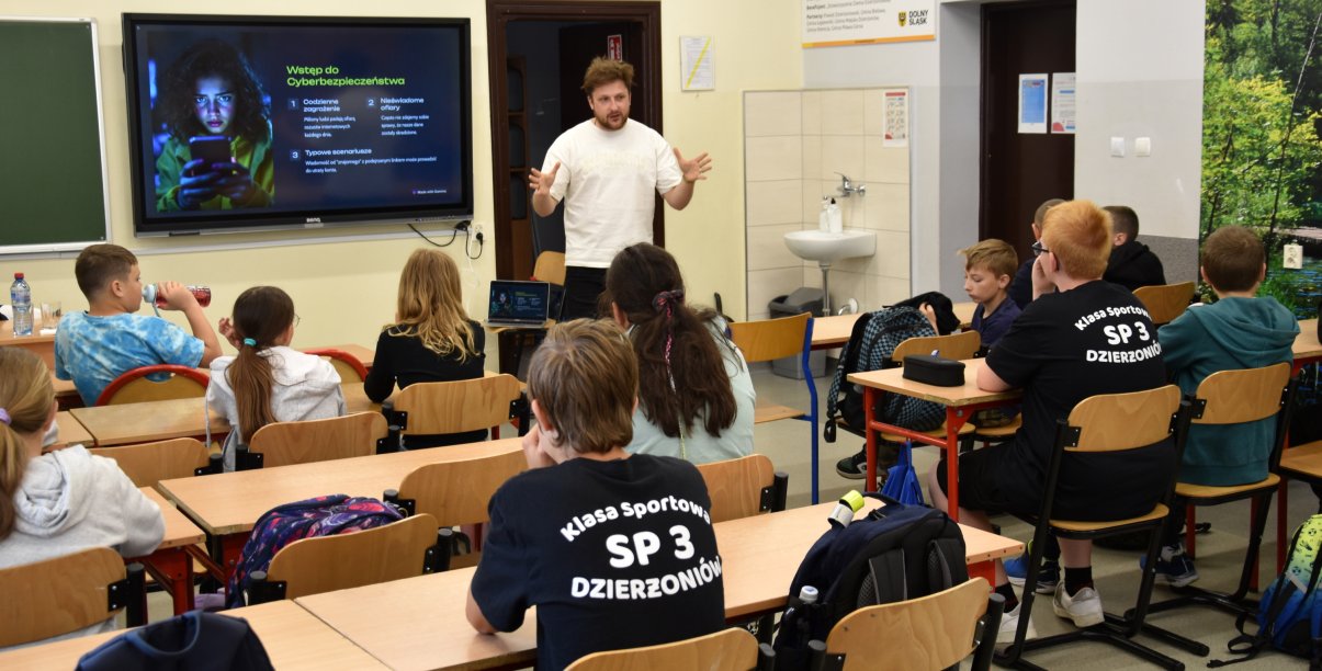 Uczniowie siedzący w ławkach szkolnych, przy interaktywnej tablicy dorosła osoba w trakcie prezentacji