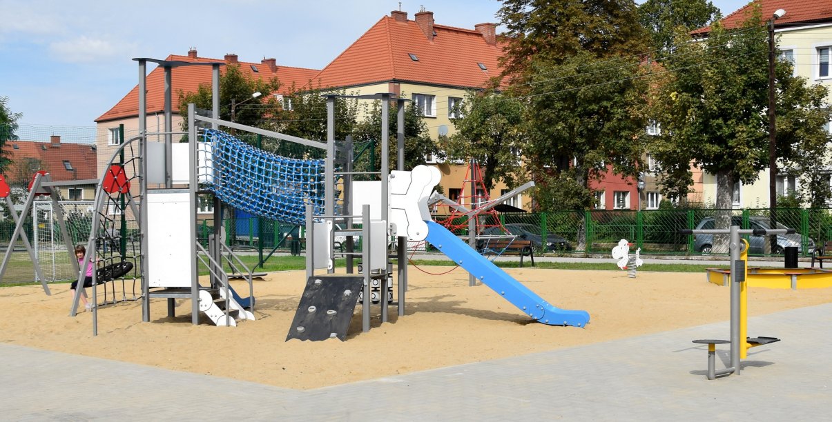 Plac z urządzeniami do zabawy zamontowanymi na piaszczystym podłożu, w tle budynki wielorodzinne