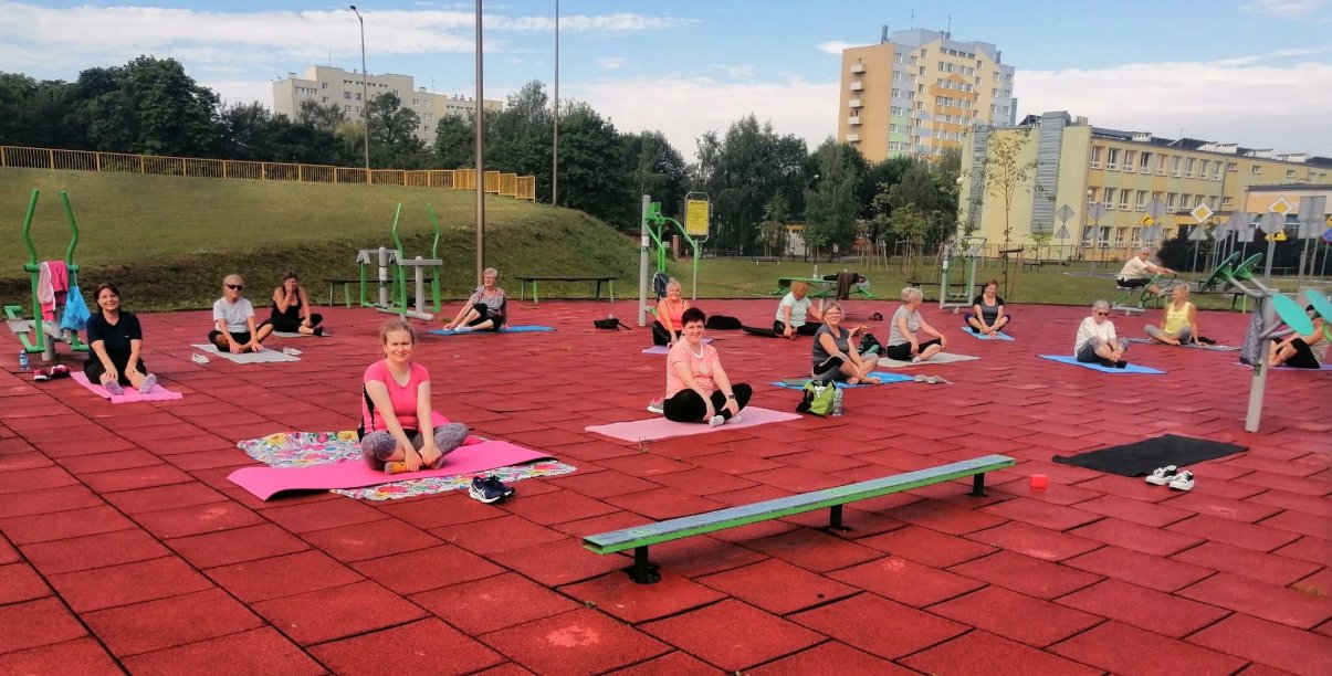 Kilkanaście osób siedzących na matach do ćwiczeń na terenie zewnętrzej siłowni, za nimi zieleń i bloki osiedlowe