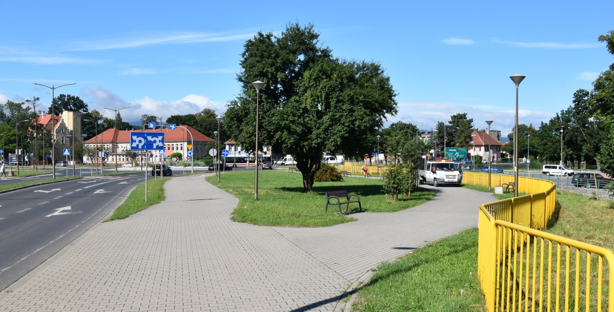 Droga w mieście, obok chodnik i teren zielony z dużym drzewem, po prawej żółte barierki