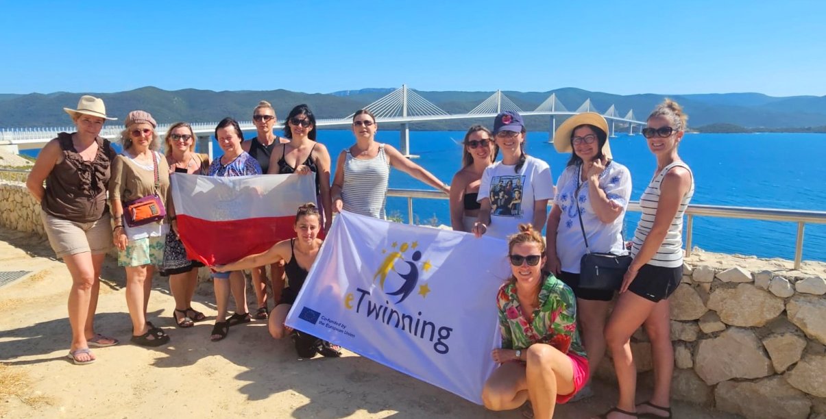 Nauczycielki z flagami Polski i projektu Twinning na tle morza, w dalekim planie duży most i góry