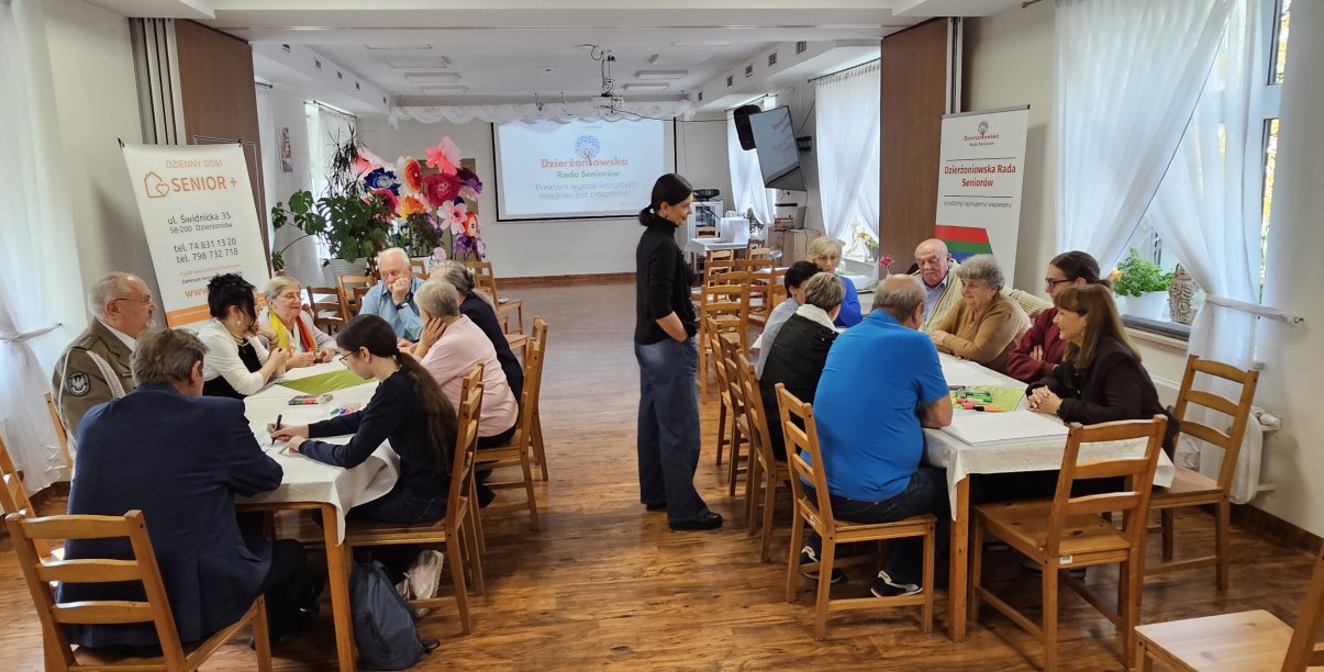 Dwie grupy ludzi przy stołach w sali multimedialnej w Centrum Seniora, pośrodku prowadząca warsztaty pani psycholog