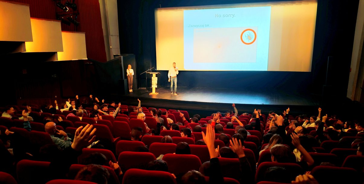 Sala Kinoteatru "Zbyszek", na scenie prowadzący wykład, nad fotelami kinowymi widać ręce zgłaszających się dzieci