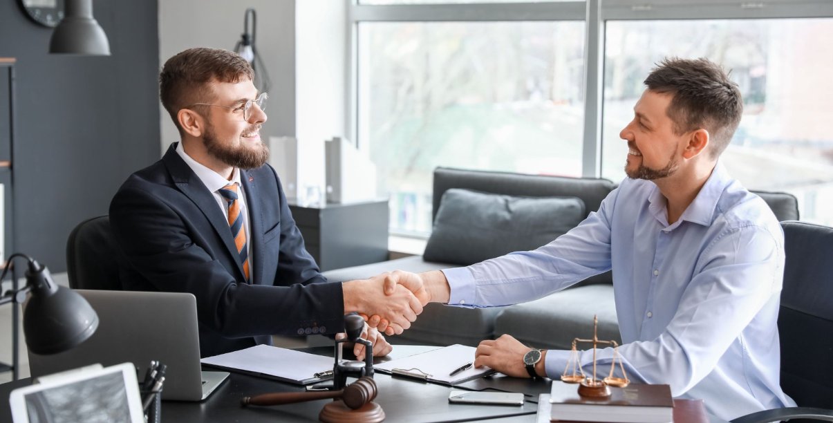 Mężczyzna w garniturze przy biurku, z dokumentami, tłumaczący coś drugiej osobie. Podają sobie wzajemnie dłoń.