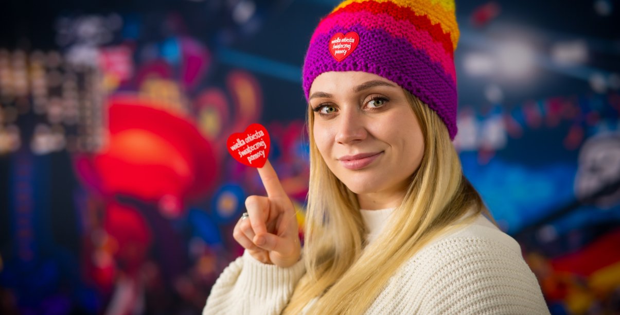 Blondynka w kolorowej czapce s zerduszkiem WOŚP trzyma na palcu serduszko WOŚP