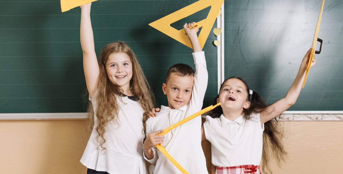 troje dzieci z przyrządami do geometrii stoi przed tablicą w klasie