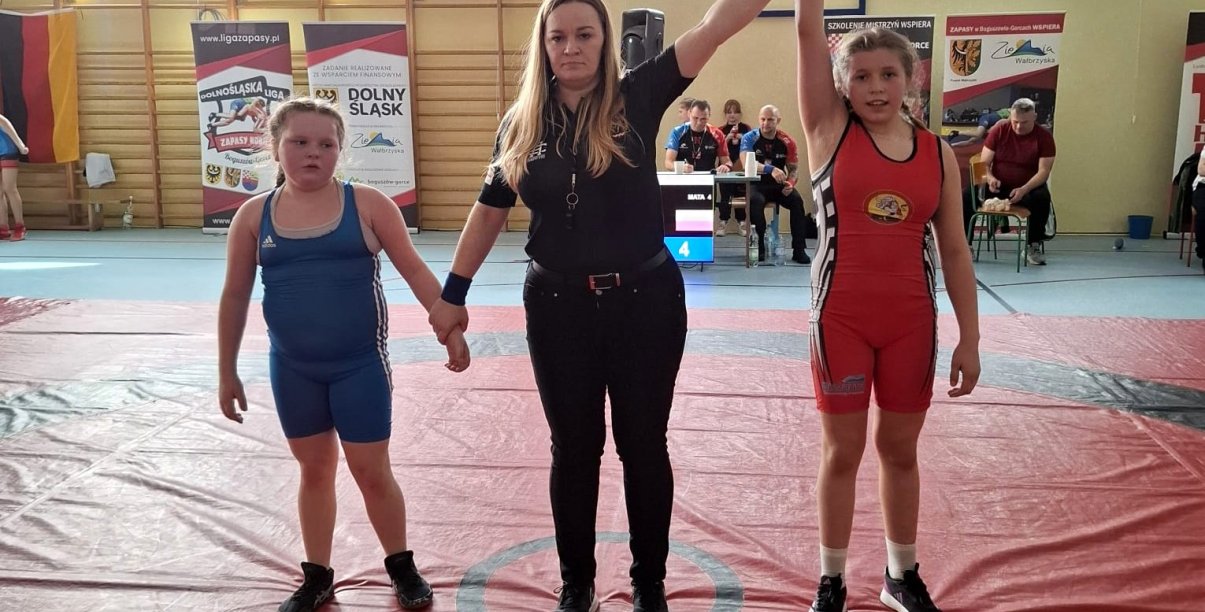 Dwie zawodniczki i sędzina na macie podczas ogłaszania wyniku