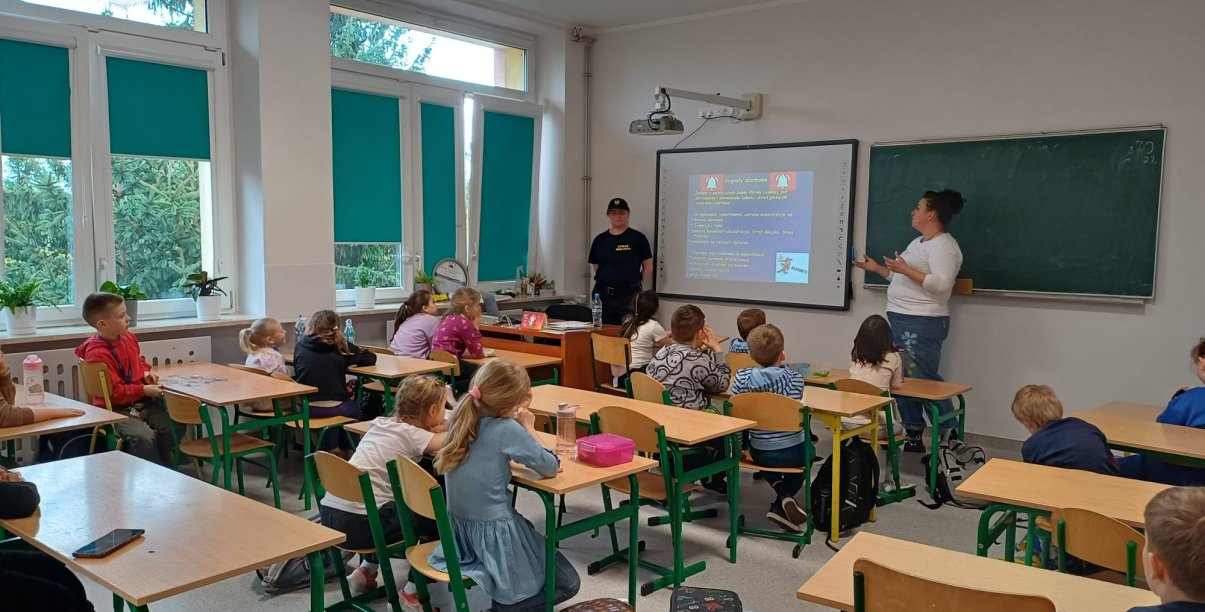 Sala w szkole, dzieci siedzą w ławkach, patrzą w kierunku tablicy, przy niej dwie kobiety, jedna z nich wskazuje ręką na tablicę