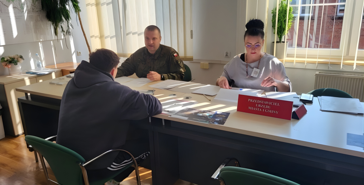 Żołnierz w mundurze i kobieta siedzący przy stole, przed nimi młody człowiek w bluzie z kapturem