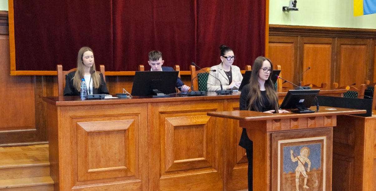 Młodzieżowi radni, dziewczyna stojąca przy mównicy, chłopak i dwie dziewczyny siedzący za stołem prezydialnym
