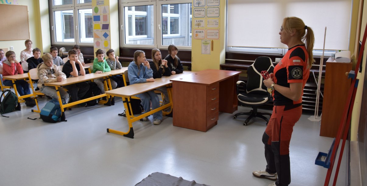 Pracownik oddziału ratunkowego w pomarańczowo-czarnym stroju stoi w klasie przed uczniami siedzącymi w ławkach