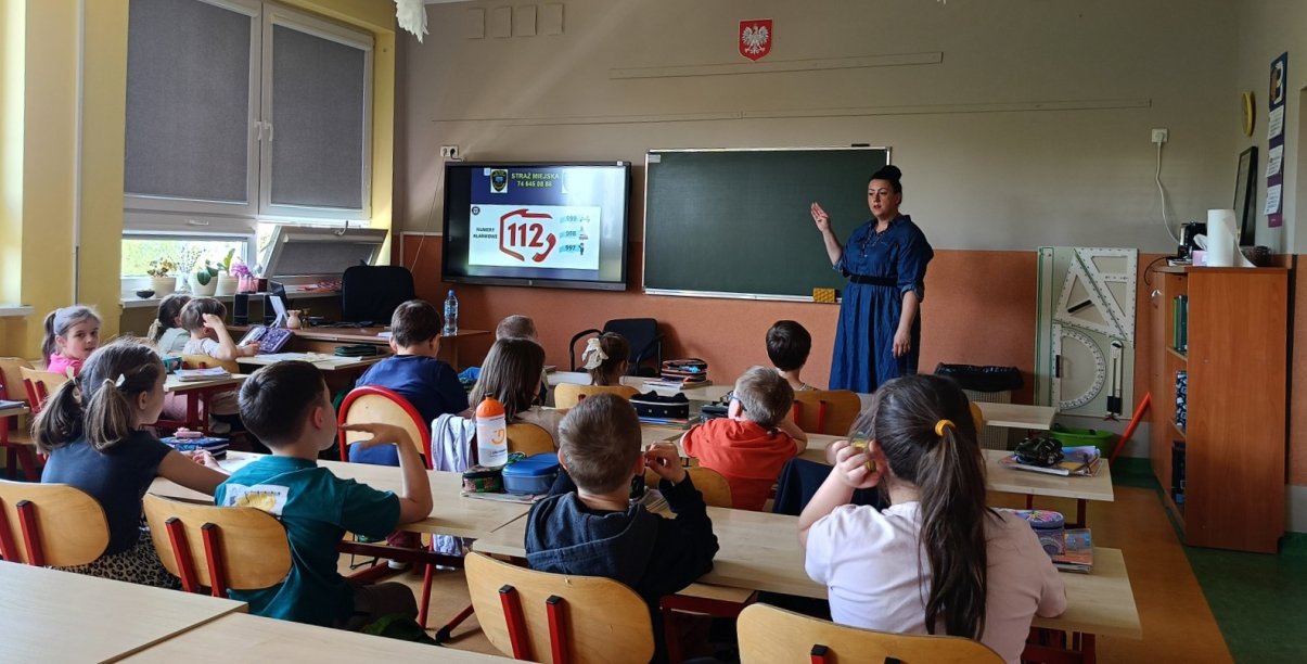 Klasa szkolna, młodzi uczniowie przy ławkach, przy tablicy pani w niebieskiej sukience