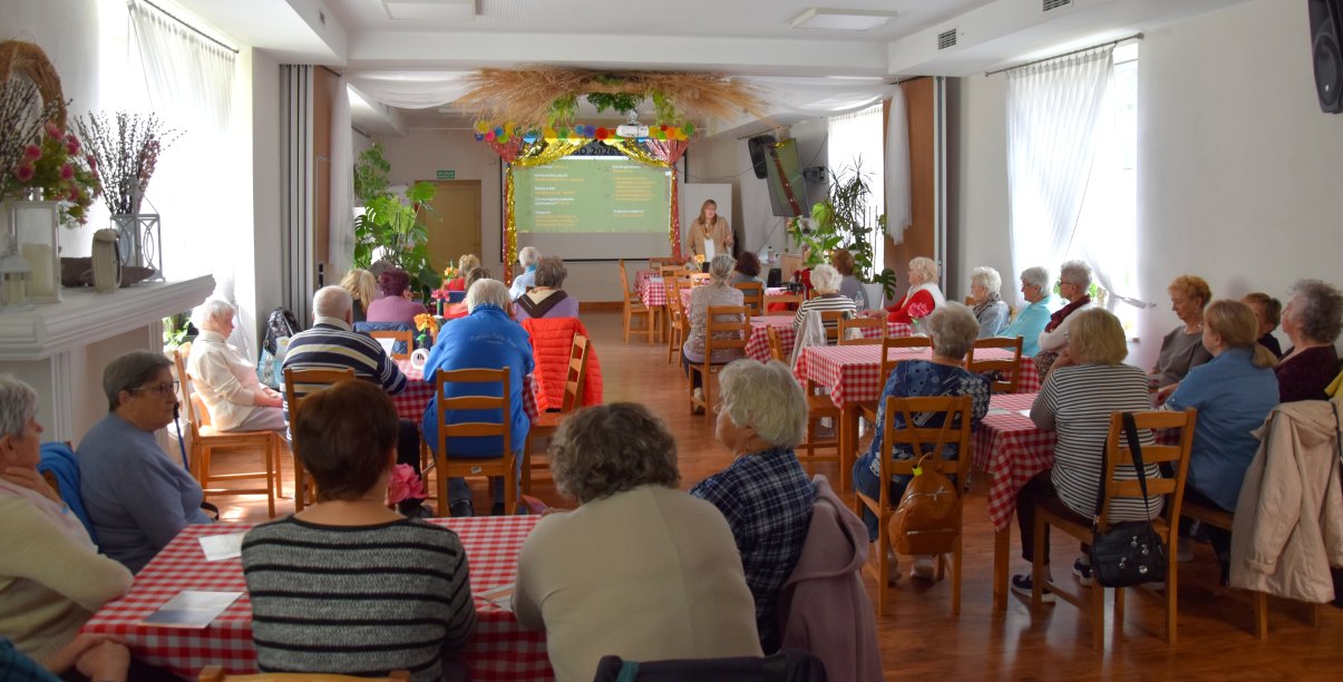 Duża sala, przy stołach siedzą seniorzy, na końcu sali kobieta przy dużym ekranie