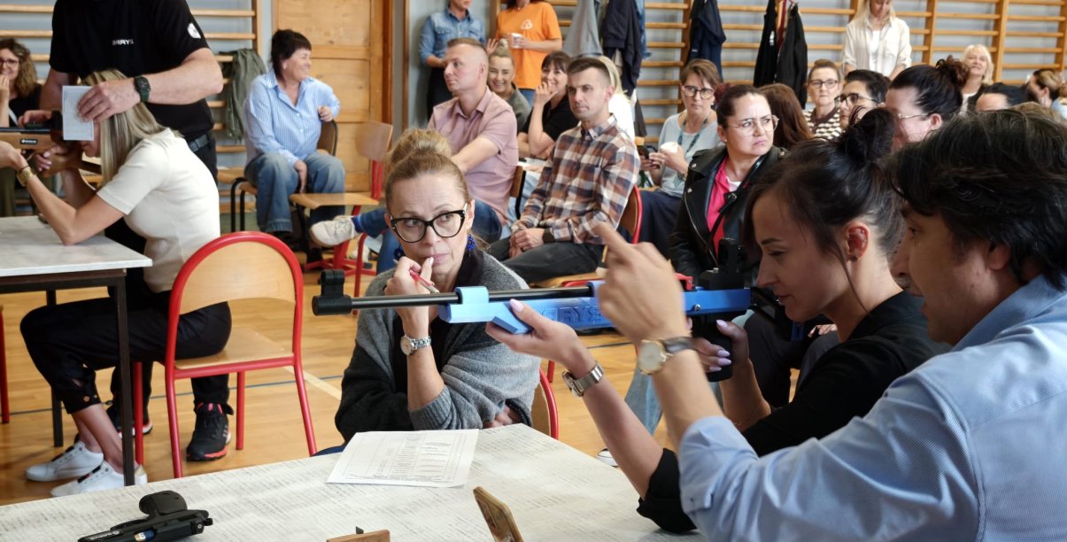 Szkolenie, wypełniona sala gimnastyczna, na pierwszym planie instruktor i kobieta trzymająca karabin