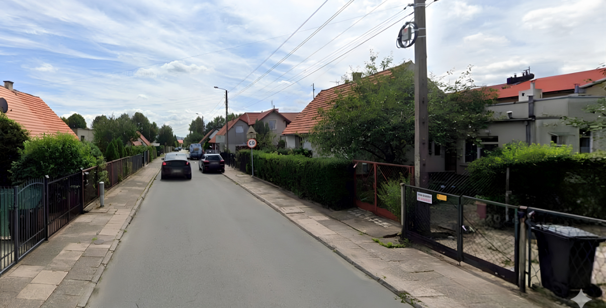 Uliczka, zaparkowane i jadące samochody, po bokach małe domki jednorodzinne