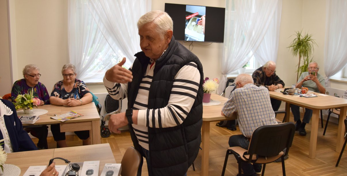 Senior z opaską na ręce, za nim przy stołach siedzi grupa seniorek i seniorów
