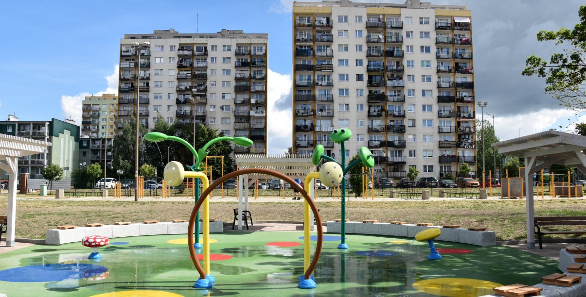 Wodny plac zabaw z zielona nawierzchnią i z urządzeniami tryskającymi wodę, w tle dwa wieżowce