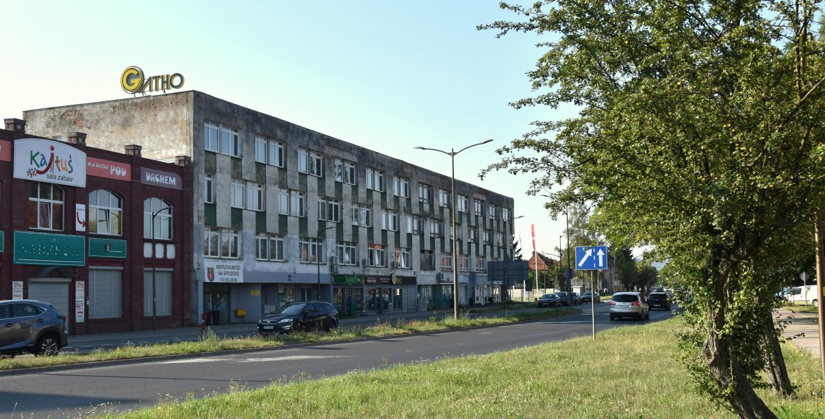 Długi trzypiętrowy budynek położony przy drodze, po prawej stronie drzewo