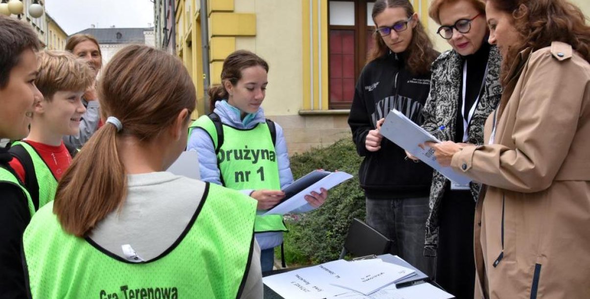 Uczniowie w zielonych kamizelkach z napisem "Gra terenowa" przy stolu ustawionym na ulicy, obok trójka organizatorów konkursu