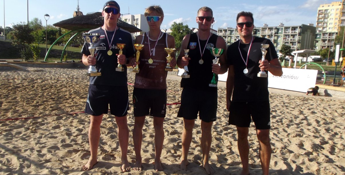 Czterech zawodników, każdy z pucharem i medalem, stojących na boisku do siatkówki plażowej, za nimi bloki osiedlowe