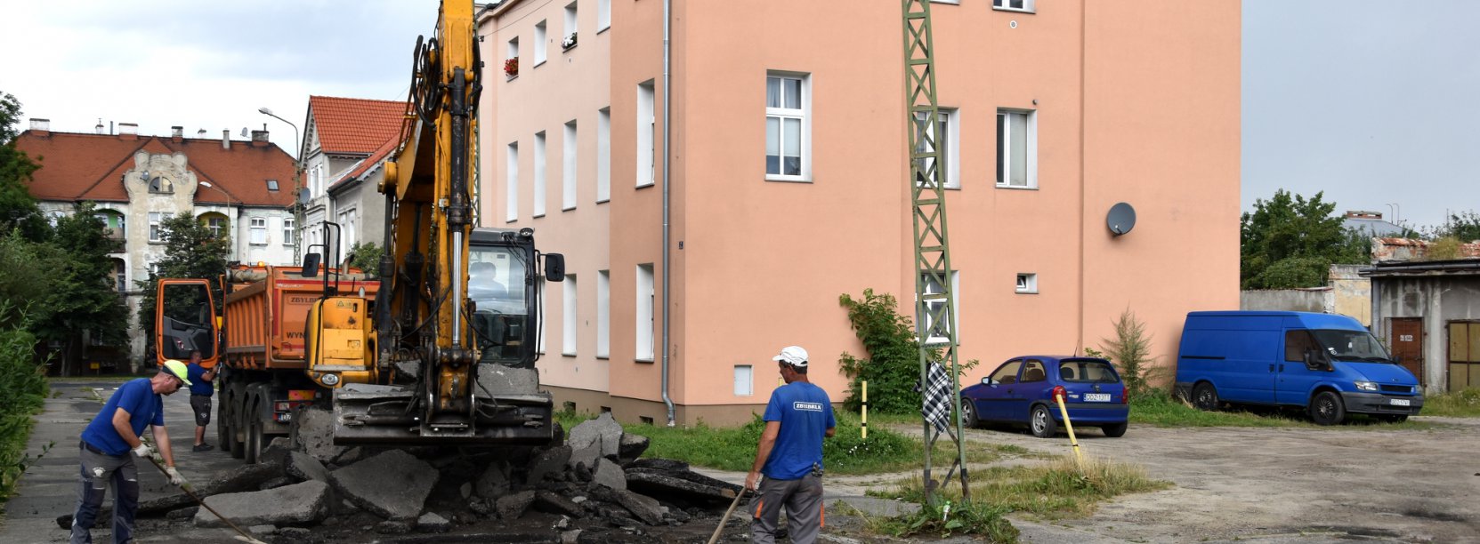 Koparka pracująća na remontowanej drodze, przy koparce pracownik, w tle budynek