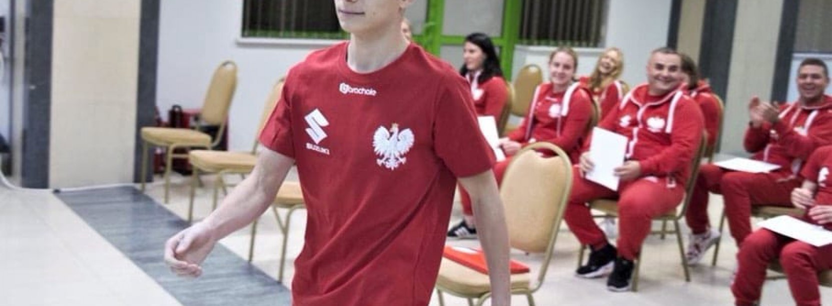 Młody zawodnik idący w koszulce z godłem, w tle gupa siedzących osób