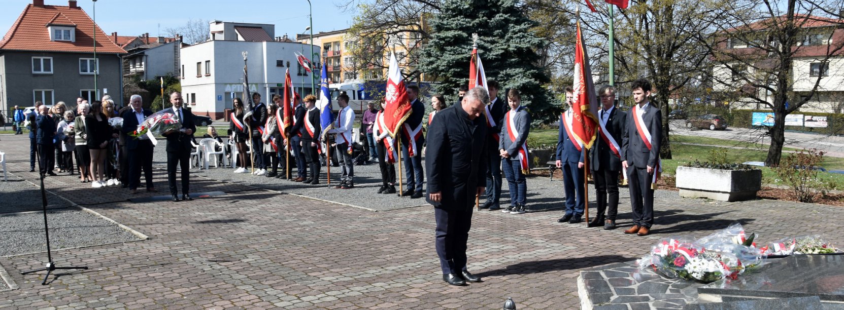 Mężczyzna stojący przed pomnikiem, w durim tle poczet sztandarowy