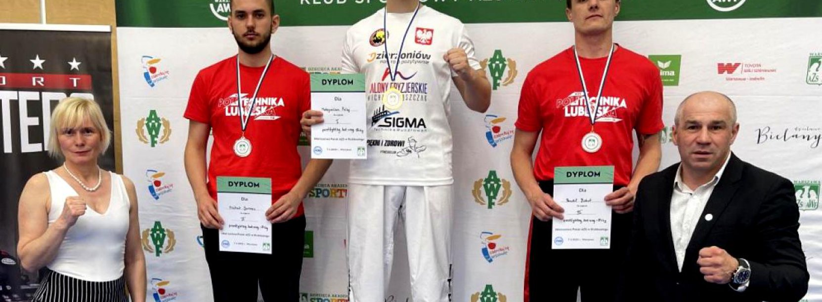 Trójka zawodników stojąca na podium, obok podium dwie osoby