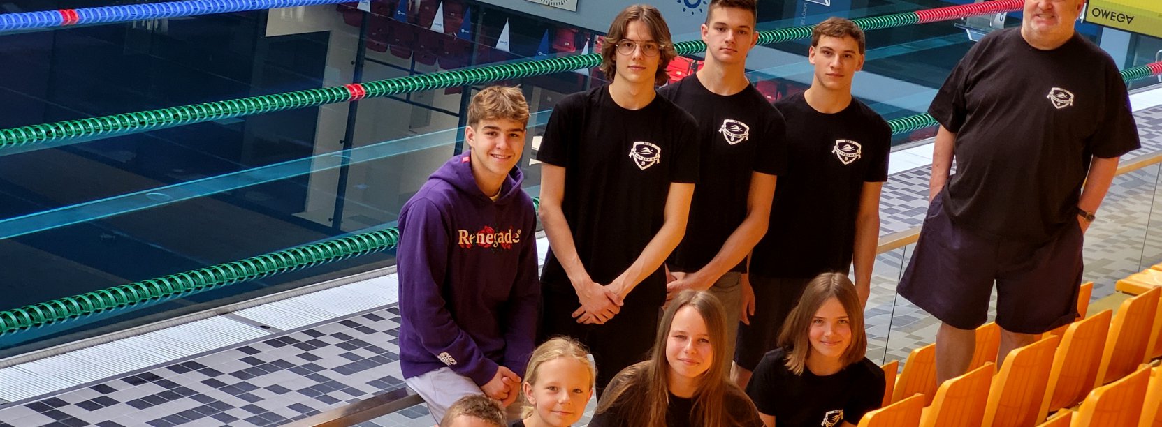 Grupowe zdjęcie młodych pływaków przy basenie, obok usmiechnięty trener