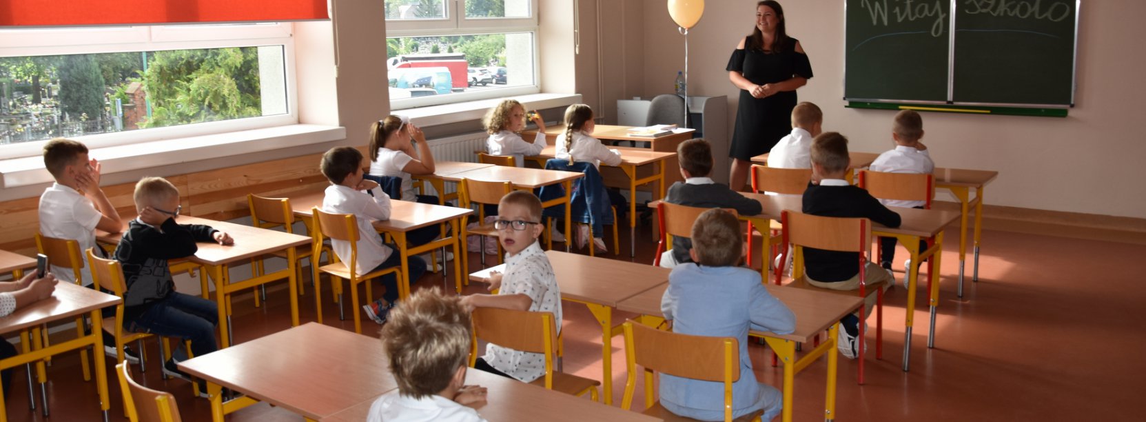 Szkolna klasa, uczniowie siedzący przy łąwkach i uśmiechnięty nauczyciel