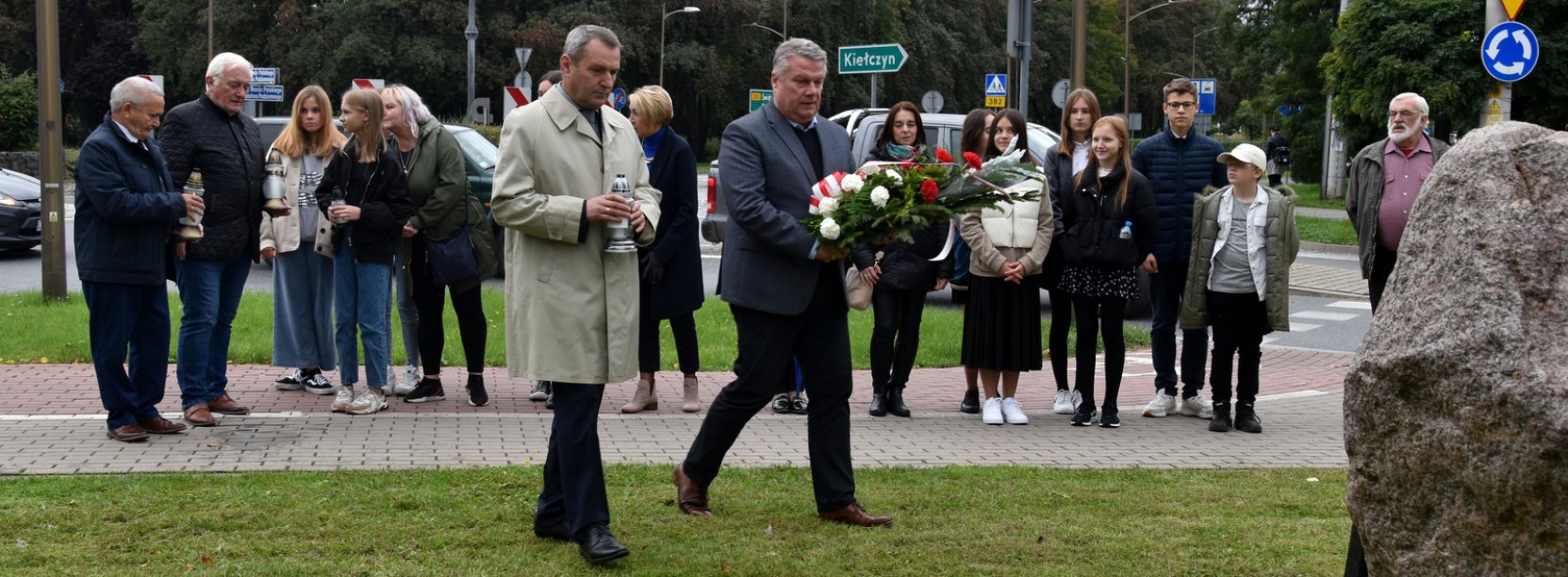 Grupa stojących osób, dwóch panów ze zniczem i kwiatami idą w kierunku kamiennego obelisku