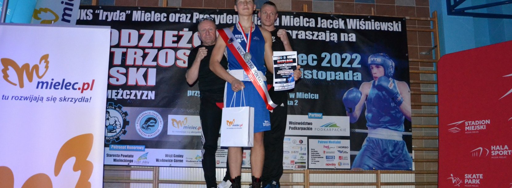 Zawodnik stojący na podium, za nim dwóch trenerów