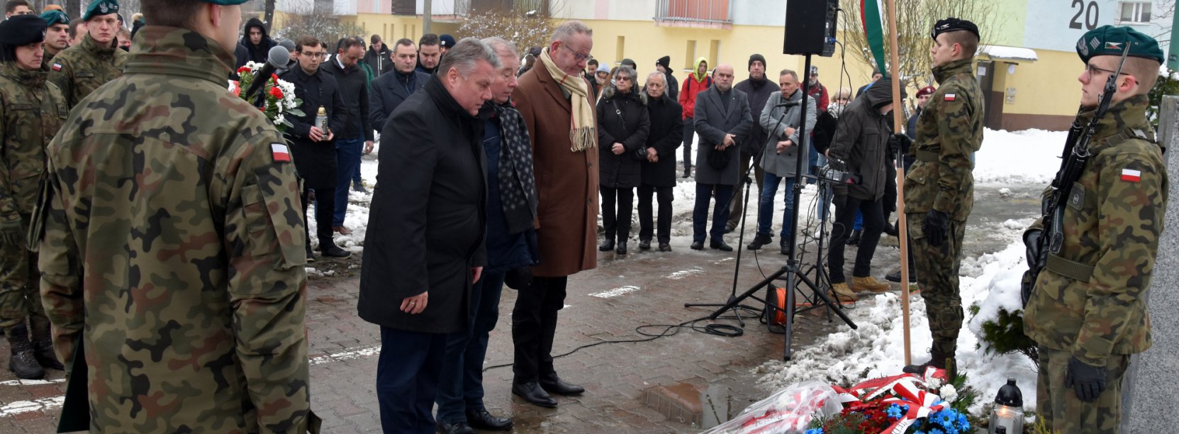 Liczna grupa osób, wśród nich żołnieże, trzy osoby składają ukłon przed obeliskiem