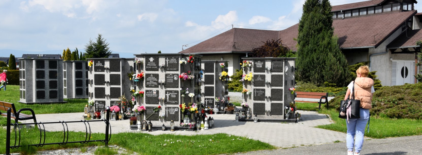 Kobieta stojąca przy kolumbarium, w drugim tle budynek kaplicy cmentarnej