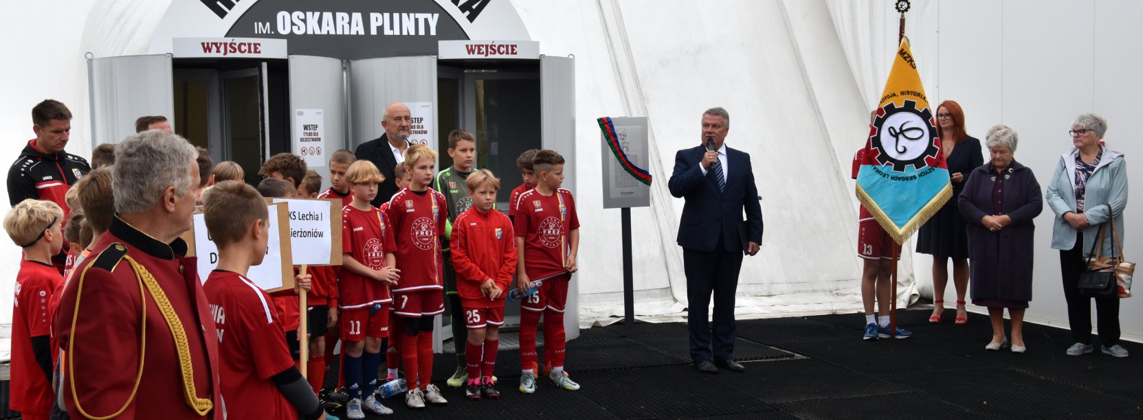 Grupa piłkarzy w czerwonych strojach przed halą piłkarską, obok mężczyzna w garniturze z mikrofonem
