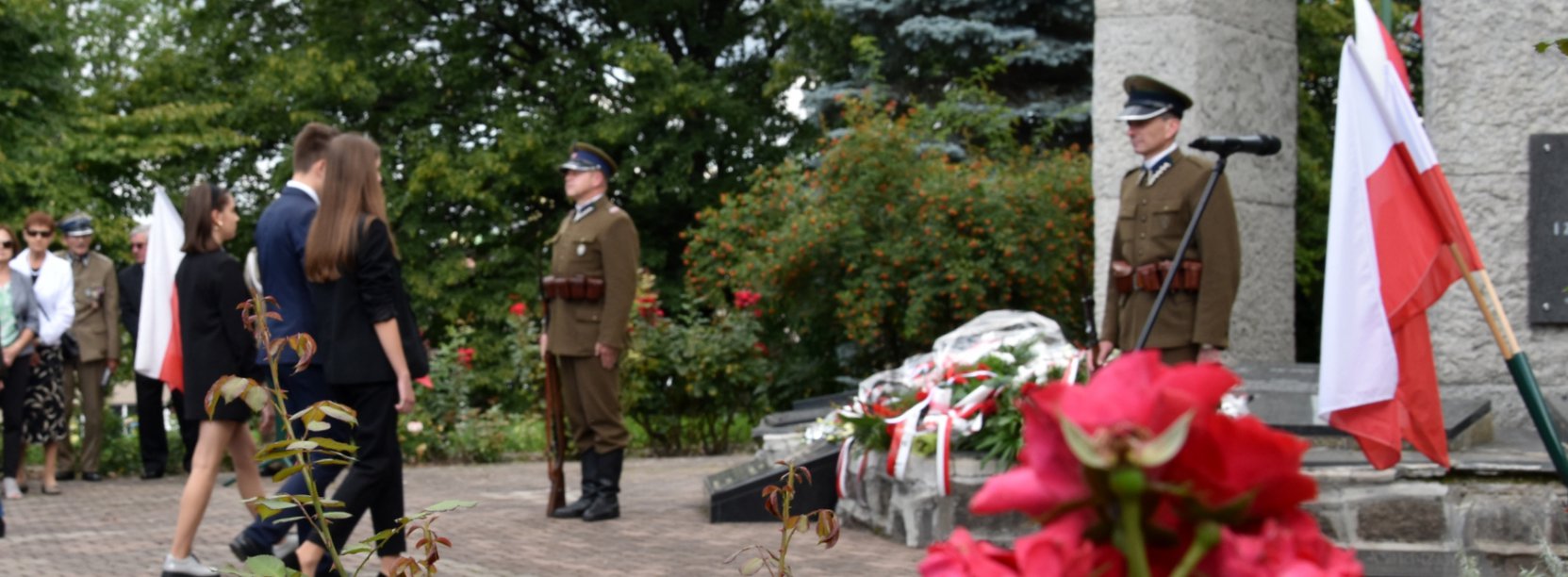 Dwóch żołnieży pełni wartę przy pomniku, do któego podchodzi grupa młodzieży z kwiatami, obok flaga Polski
