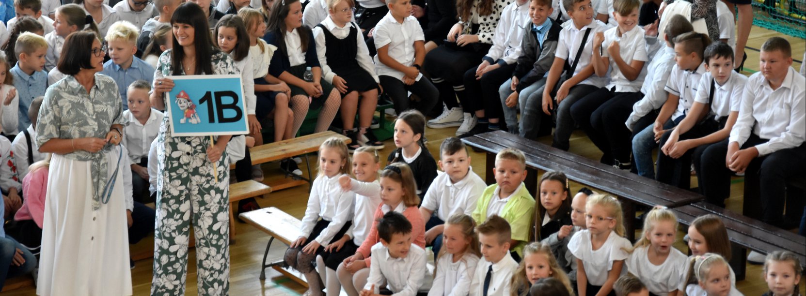 Grupowie zdjęcie młodych uczniów siedzących na ławkach, obok nauczycielka z tabliczką z napisem Klasa 1B