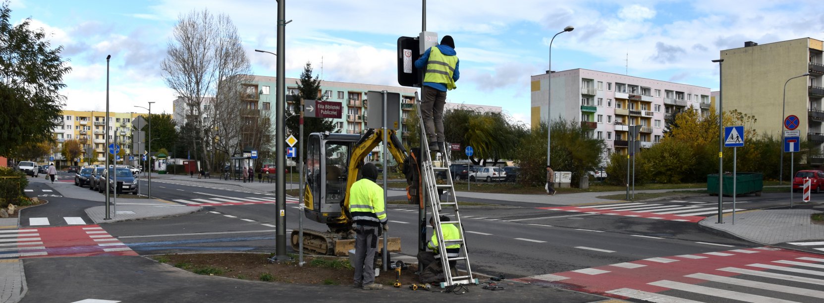 Trzy osby w jaskrawych kamizelkach montuje znak przy skrzywoniu dróg, w tle bloki mieszkalne