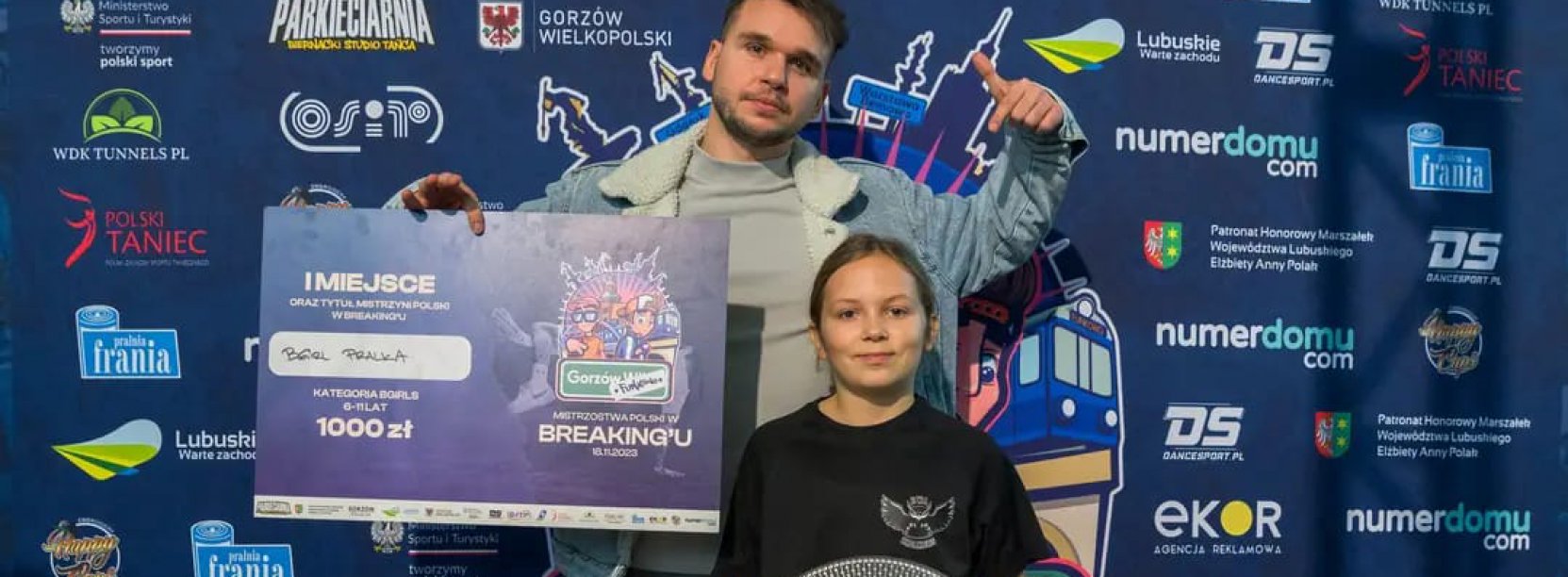 Trener trzyma w reku duży czek, przed nim zawodniczka ze srebrną paterą w tle ścianka promocyjna