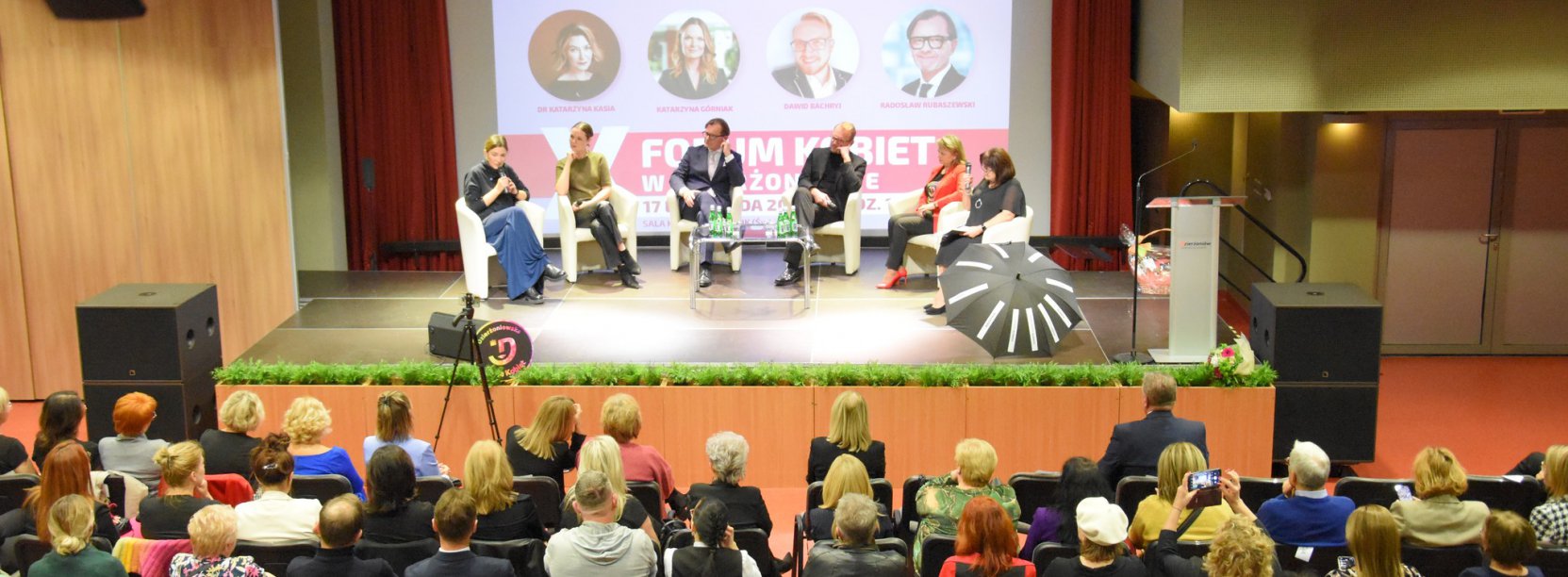 Sala, na scenie osoby siedzące w fotelach, przed nimi publiczność