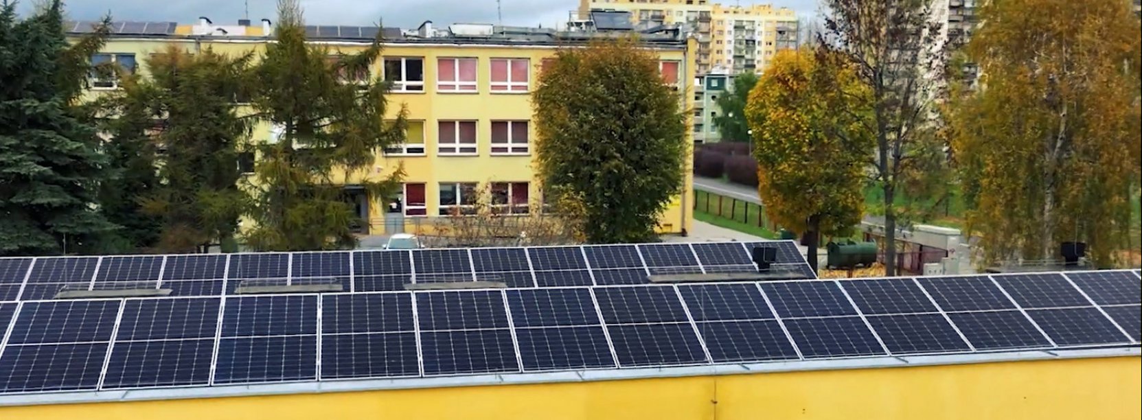 Panele fotowoltaiczne na dachu budynku, w tle osiedle mieszkaniowe