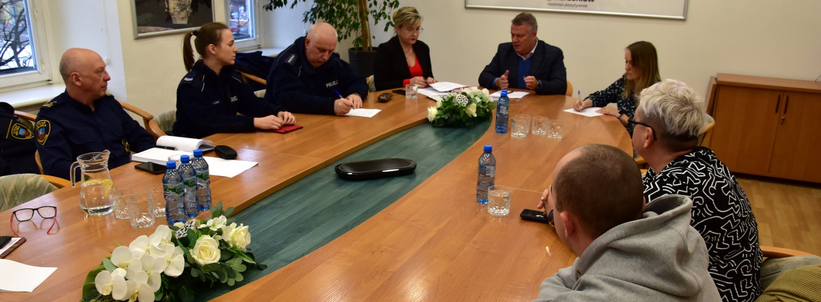 Strażnik miejski, policjanci oraz kobiety i mężczyźni po cywilnemu siedzący przy dużym owalnym stole