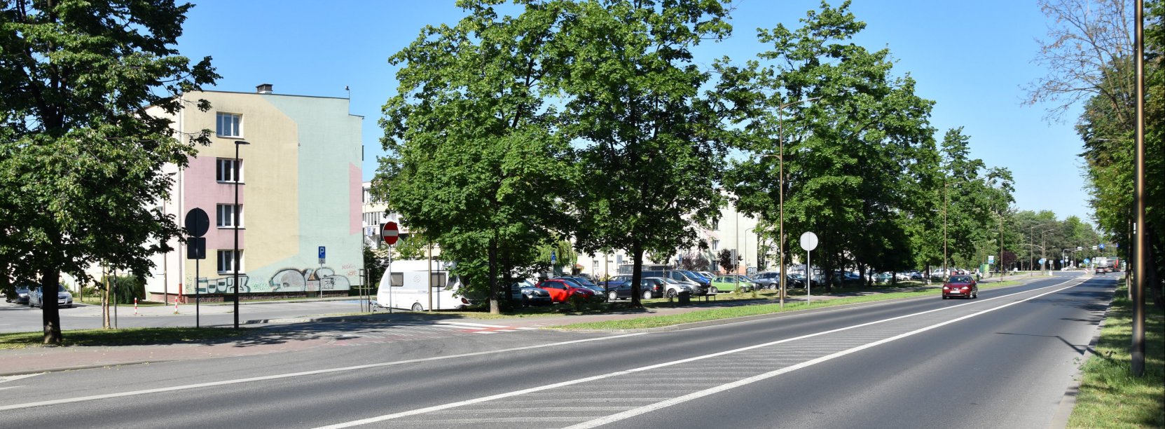 Dwupasmowa droga, wzdłuż drogi duże drzewa, za nimi bloki osiedlowe