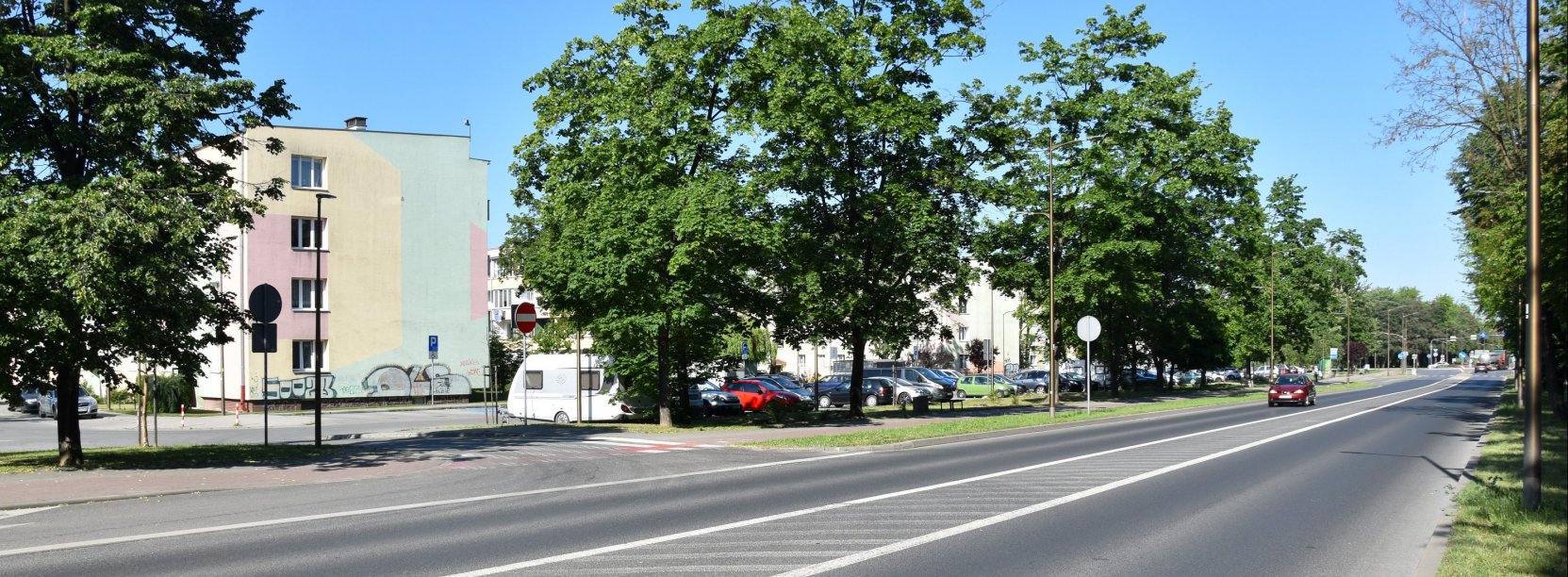 Szeroka drga przedzielona białymi pasami, za drogą drzewa i bloki mieszkalne