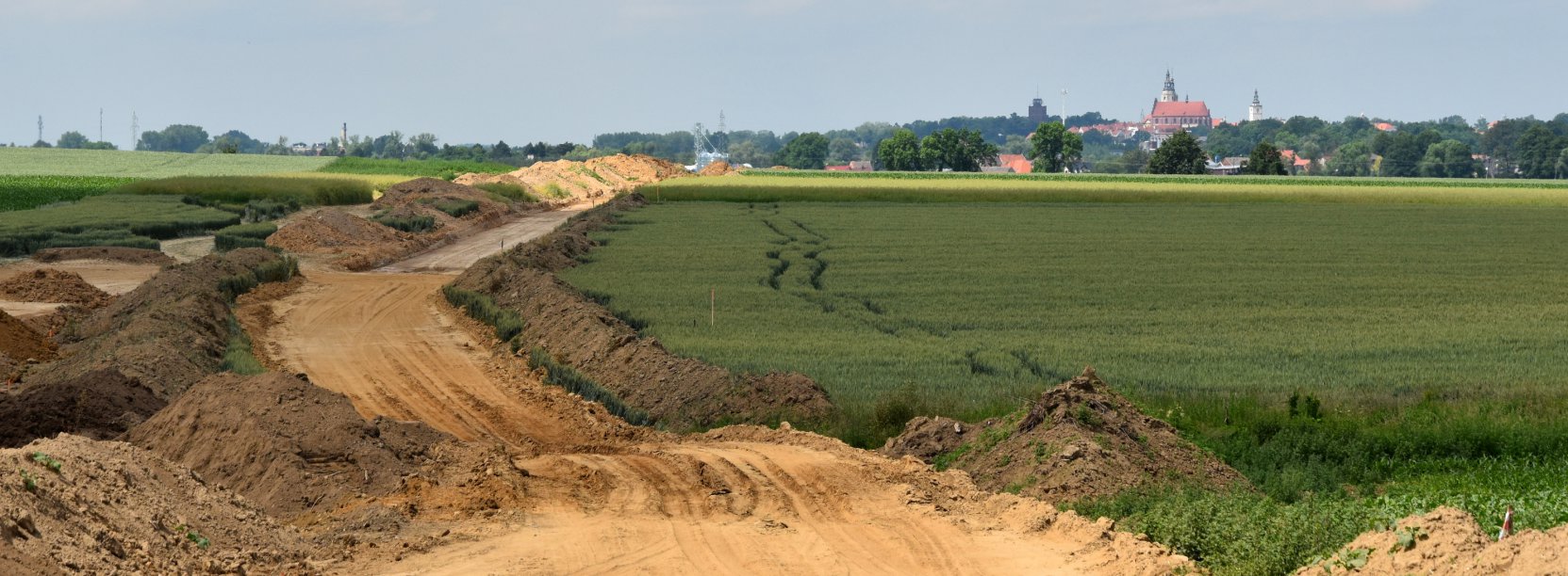 Droga w początku budowy, po bo bokach zielone pola, w dali panorama miasta